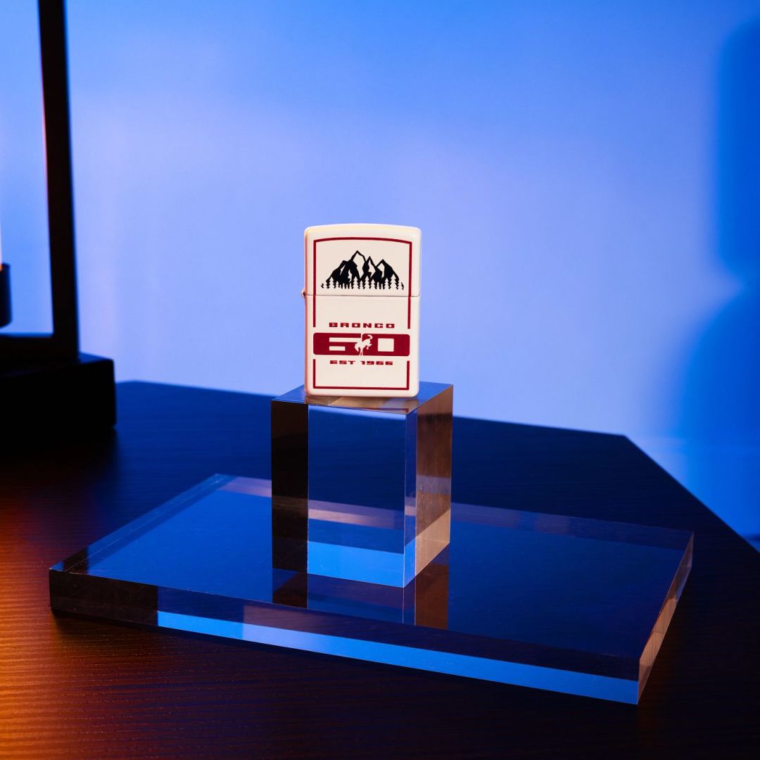 Lifestyle image of Zippo Ford Bronco 60th Anniversary Design White Matte Windproof Lighter standing on two clear blocks on a table showing the front of the lighter with a blue light showing through the clear blocks and on to the wall. 

