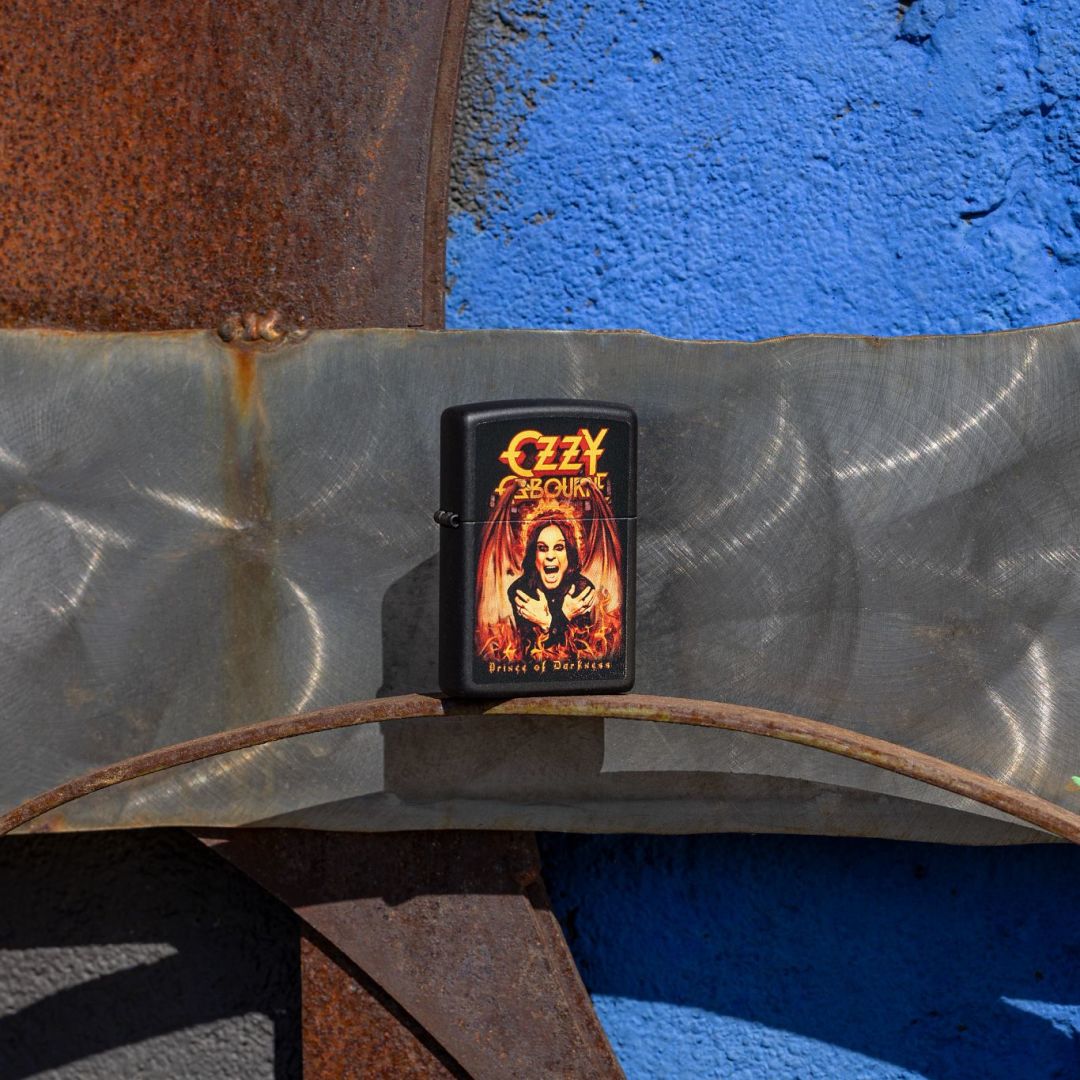 Lifestyle image of Zippo Ozzy Osbourne Prince of Darkness Design Black Matte Windproof Lighter standing on a rusty 3-D steel sculpture facade showing the front and hinge side of the lighter with a rusty metal and blue background. 

