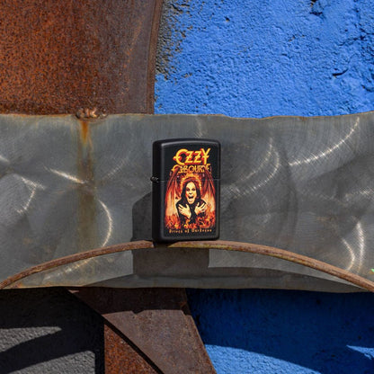 Lifestyle image of Zippo Ozzy Osbourne Prince of Darkness Design Black Matte Windproof Lighter standing on a rusty 3-D steel sculpture facade showing the front and hinge side of the lighter with a rusty metal and blue background. 

