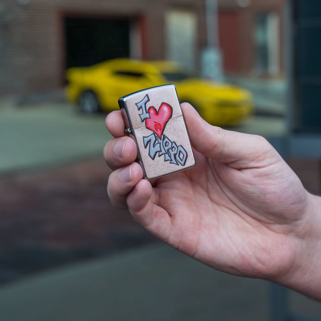 Lifestyle image of Zippo I Love Zippo Design Street Chrome Windproof Lighter in hand showing the front of the lighter with an out of focus yellow sports car parked in front of a brick building as the background.

