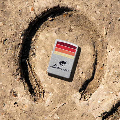 Lifestyle image of Zippo Ford Bronco Sunset Stripes Design Street Chrome Windproof Lighter lying in the middle of a horseshoe print in the mud showing the front of the lighter. 

