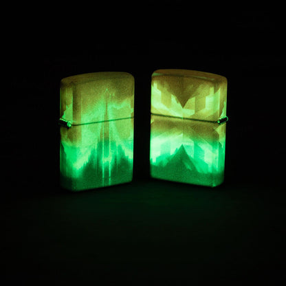 Lifestyle image of two Zippo Wicked City Design Glow in the dark Green Windproof Lighters; one showing the front of the lighter and the other showing the back and hinge side of the lighter, both glowing in the dark standing on a dark surface. 

