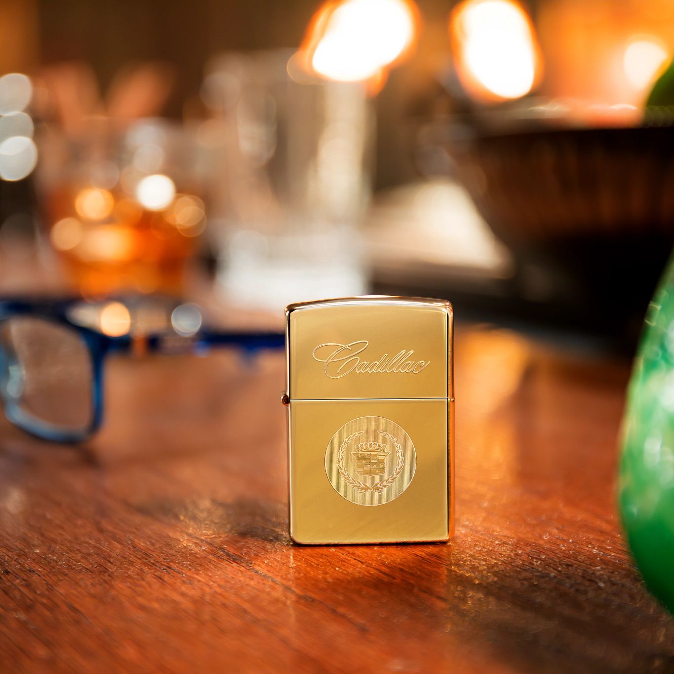 Lifestyle image of Zippo Cadillac® Crest Design High Polish Brass Windproof Lighter standing on a wooden table next to blue eyeglasses, a candle in a green glass jar, and a bowl of fruit.

