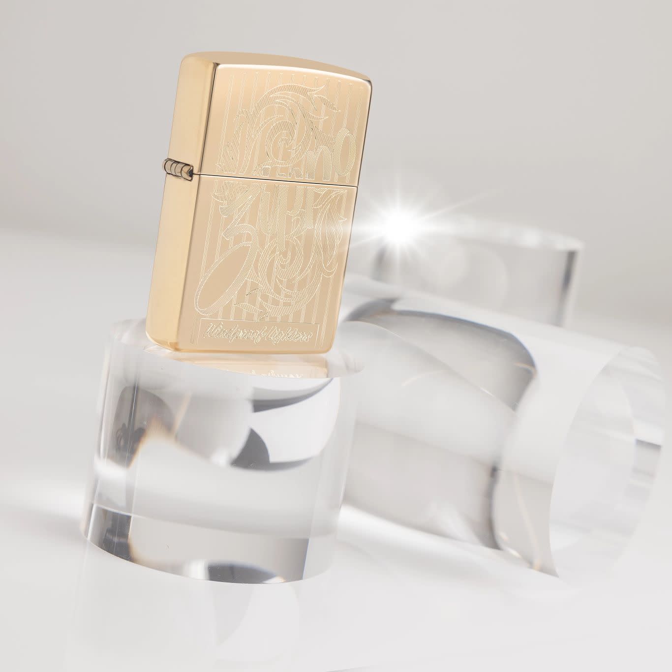 Lifestyle image of Zippo Swirling Filigree Design High Polish Brass Windproof Lighter standing on a clear cylinder with two cylinders behind it, one laying on its side.