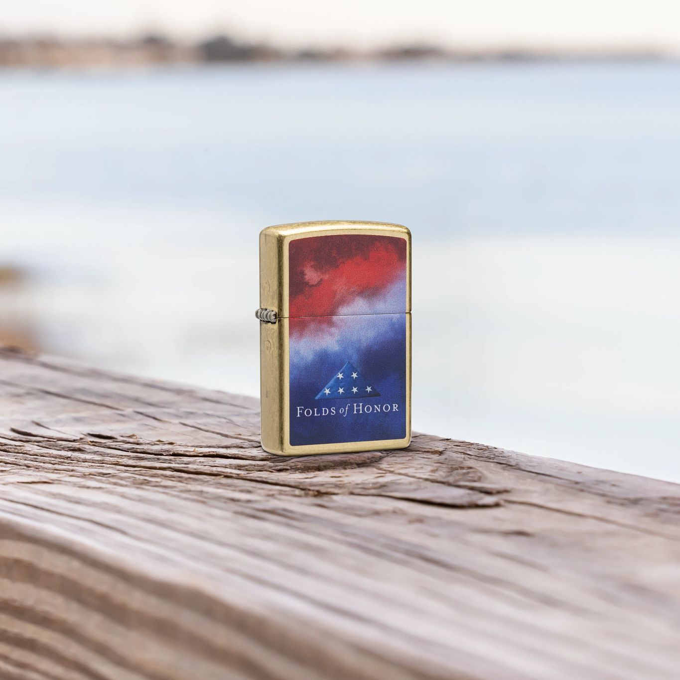 Lifestyle image of Zippo Folds of Honor Clouds Design Street Brass Windproof Lighter standing on a wooden deck railing with a body of water as the background, showing the front of the lighter. 

