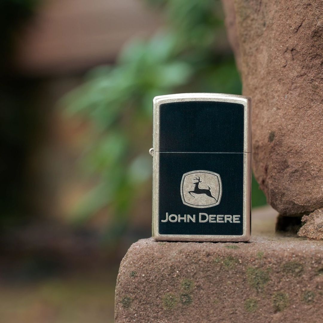 Lifestyel image of Zippo John Deere Chrome Logo Design Street Chrome Windproof Lighter standing on a rock ledge showing the front of the lighter with an out of focus nature background. 

