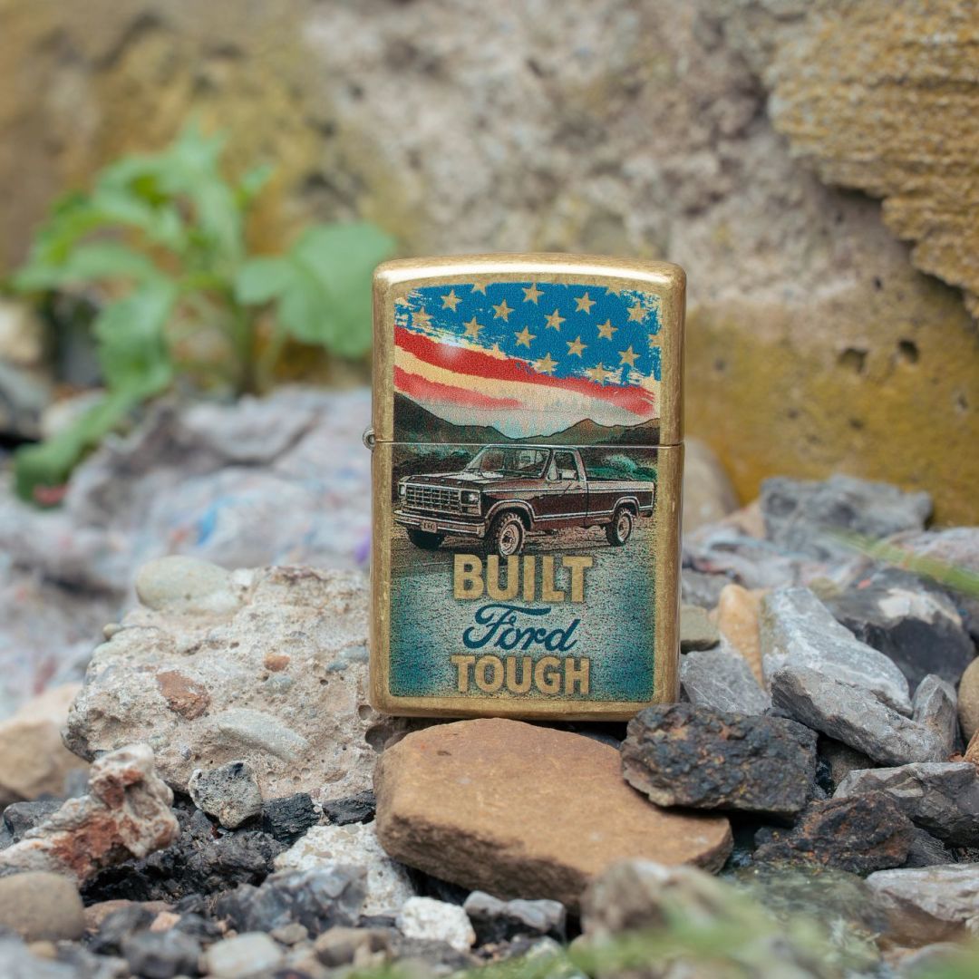 Lifestyle image of Zippo Built Ford Tough Design Street Brass Windproof Lighter standing on rocks showing the front of the lighter with rocks and some greens in the background. 

