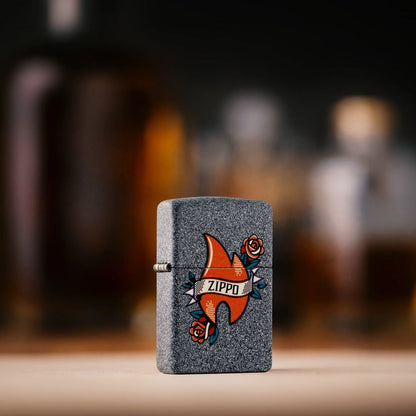Lifestyle image of Zippo Flame Tattoo Design Iron Stone Windproof Lighter standing on a light-colored countertop showing the front and hinge side of the lighter with an out of focus bar in the background. 

