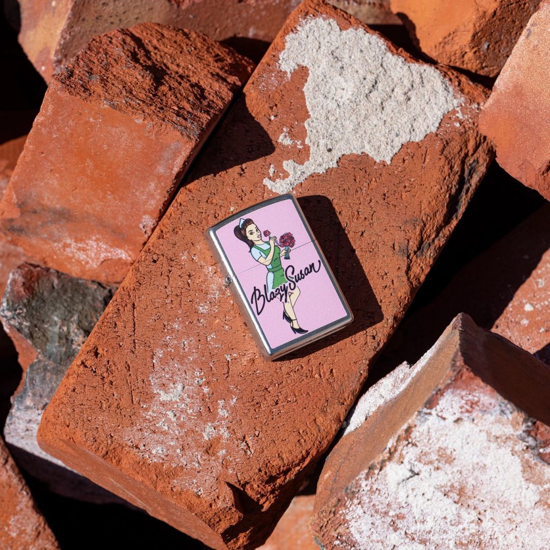 Lifestyle image of Zippo Blazy Susan Bouquet Design Street Chrome Windproof Lighter lying on a brick while showing the front of the lighter with other bricks as the background. 

