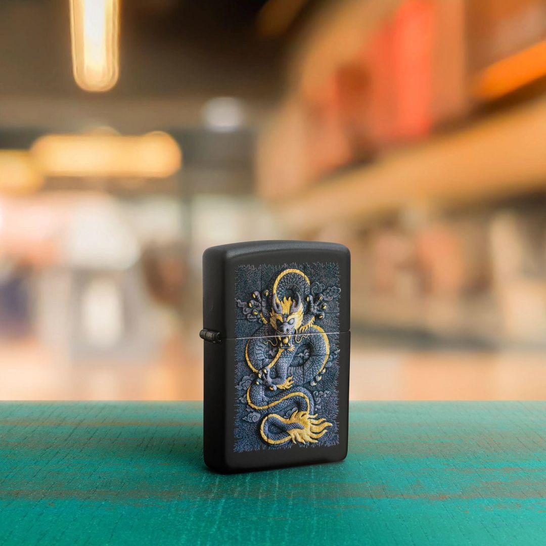 Lifestyle image of Zippo Stone Dragon Design Black Matte Windproof Lighter standing at a 3/4 angle on a teal table showing the front of the lighter with an out of focus shopping mall as the background. 

