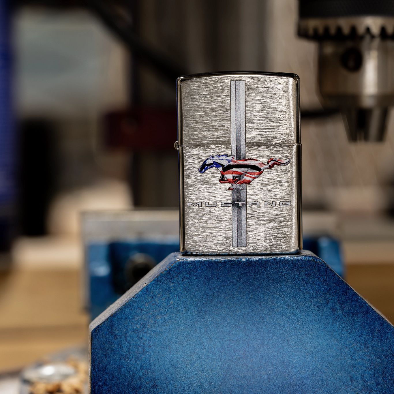 Lifestyle image of Zippo Ford Mustang Design Brushed Chrome Windproof Lighter standing on top of a heavy-duty vise grip with automotive tools in the background. 

