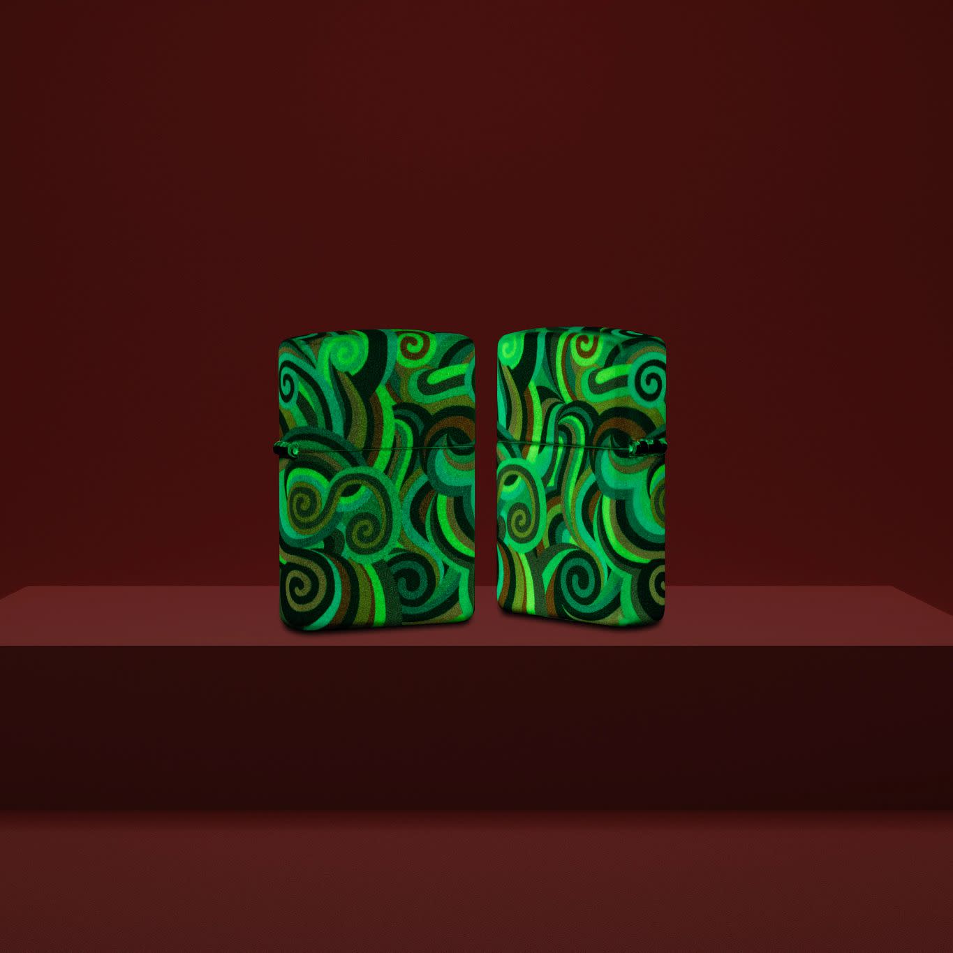 Lifestyle image of two Zippo Rick Rietveld Glowing Swirls Design Glow in the Dark Windproof Lighters standing on a burgundy pedestal glowing in the dark.