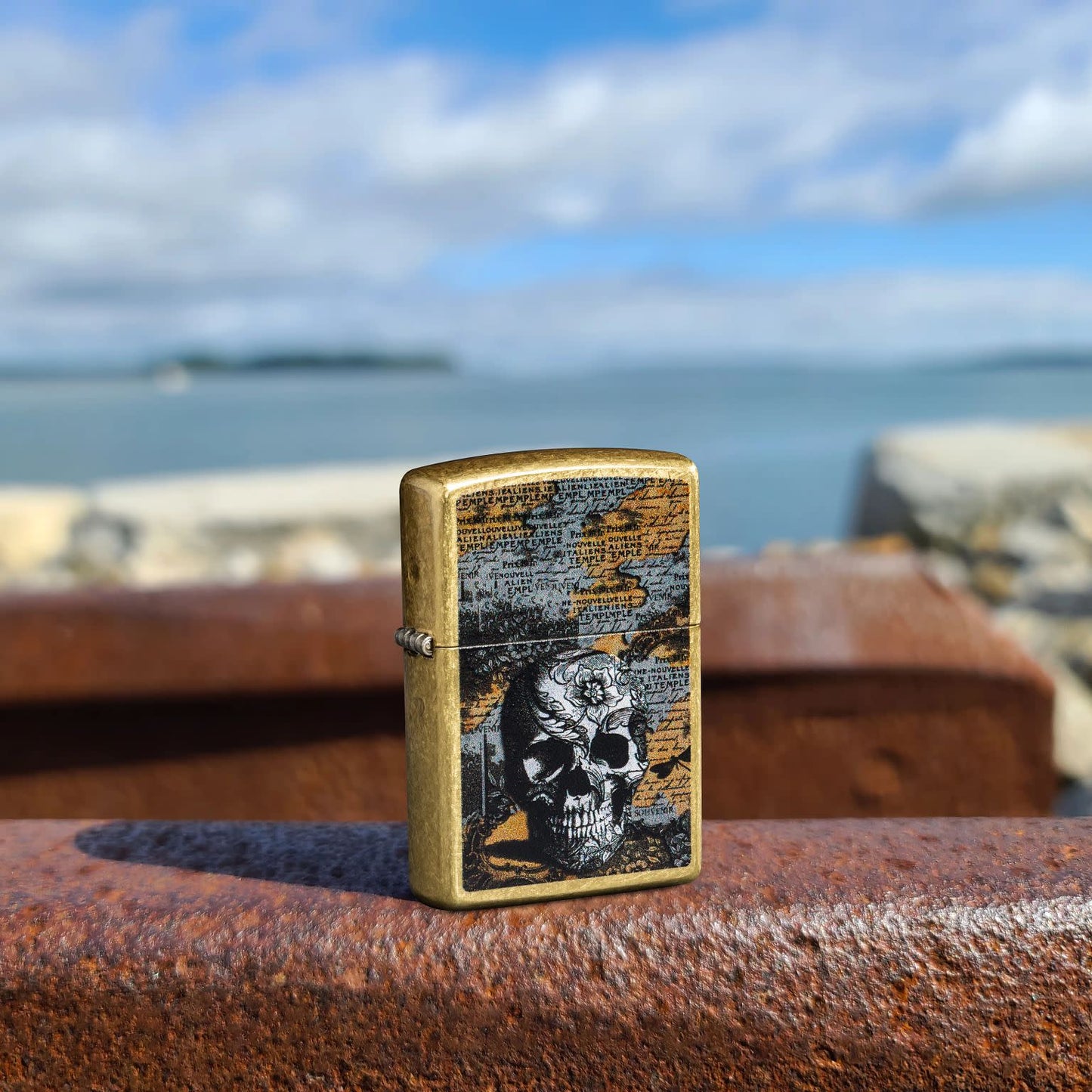 Lifestyle image of Zippo Floral Skull Design Street Brass Windproof Lighter standing on rusted machinery with a body of water and cloudy blue sky in the background.