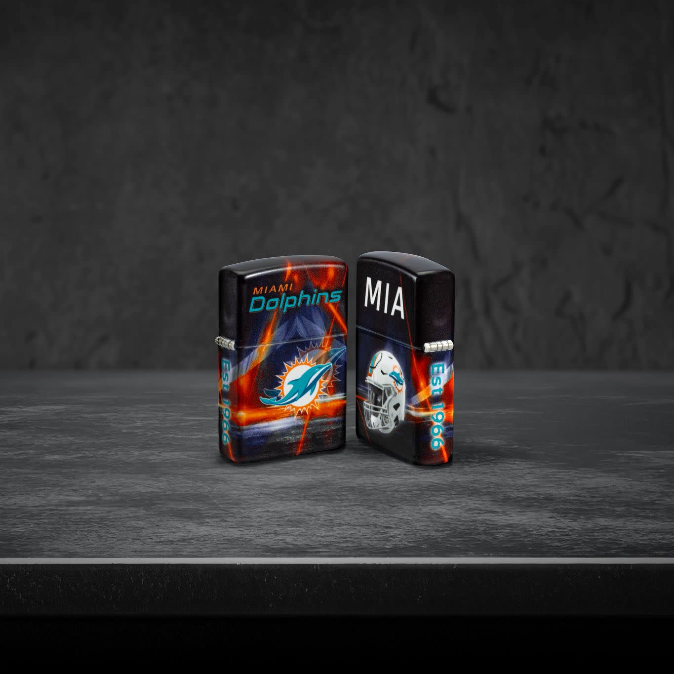 Lifestyle image of two Zippo NFL Miami Dolphins 540 Matte Windproof Lighters, one showing the front of the lighter and the other showing the back, standing on a dark grey surface.