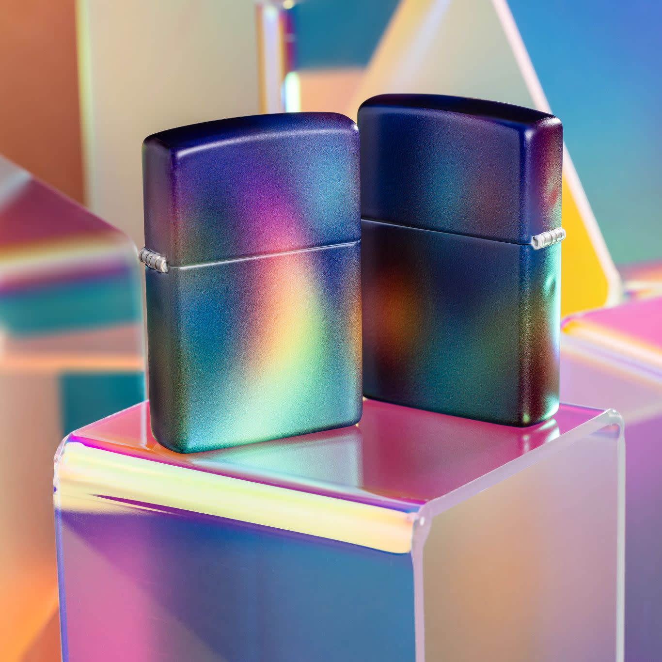 Lifestyle image of two Zippo Prismatic Design 540 Matte Windproof Lighters, one showing the front of the lighter and the other showing the back, standing on a multicolored cube with more cubes in the background.