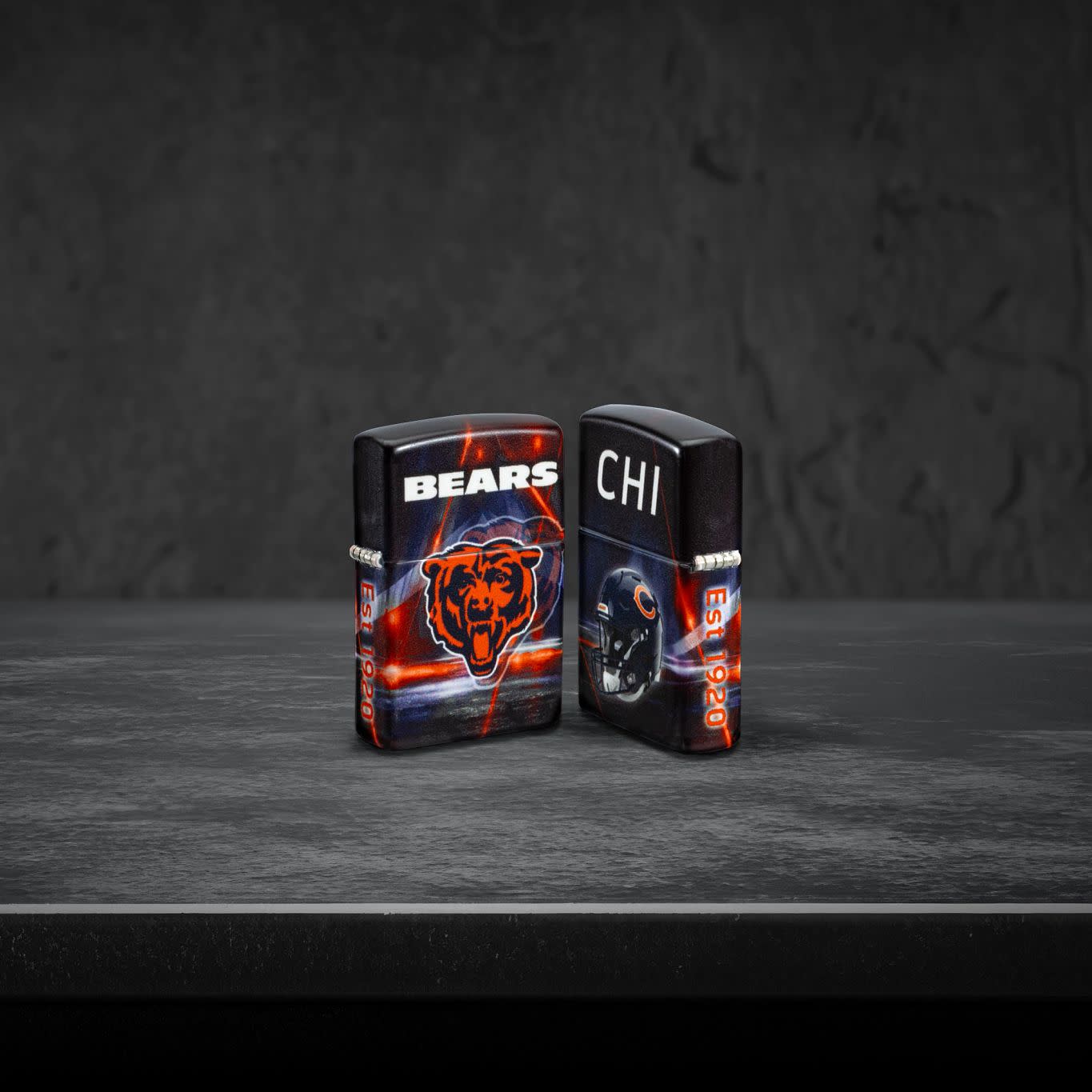 Lifestyle image of two Zippo NFL Chicago Bears 540 Matte Windproof Lighters, one showing the front of the lighter and the other showing the back, standing on a dark grey surface.