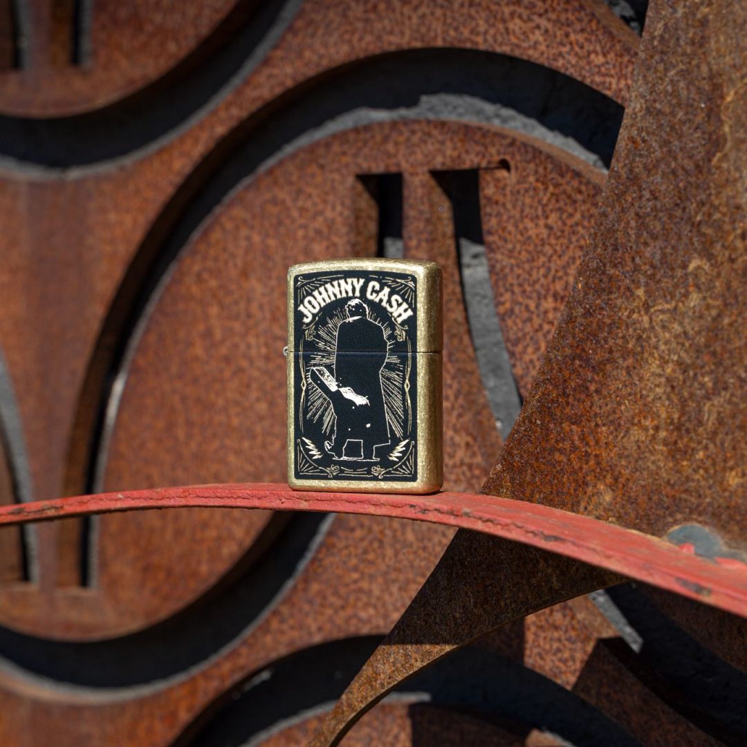 Lifestyle image of Zippo Johnny Cash Walk Design Street Brass Windproof Lighter standing in front of a rusty 3-D steel sculpture facade, showing the front of the lighter. 

