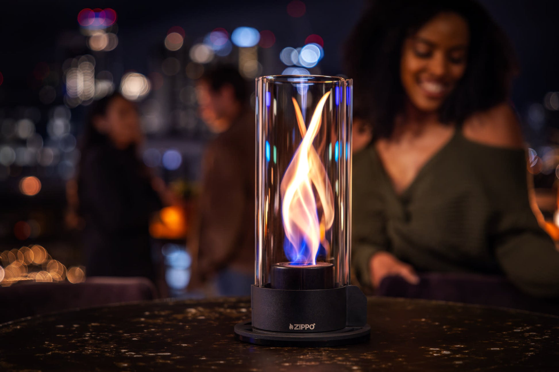 Lifestyle image of Zippo FlameScapes™ Spiral Fire Feature in black, lit on a table with a woman and couple in the background.