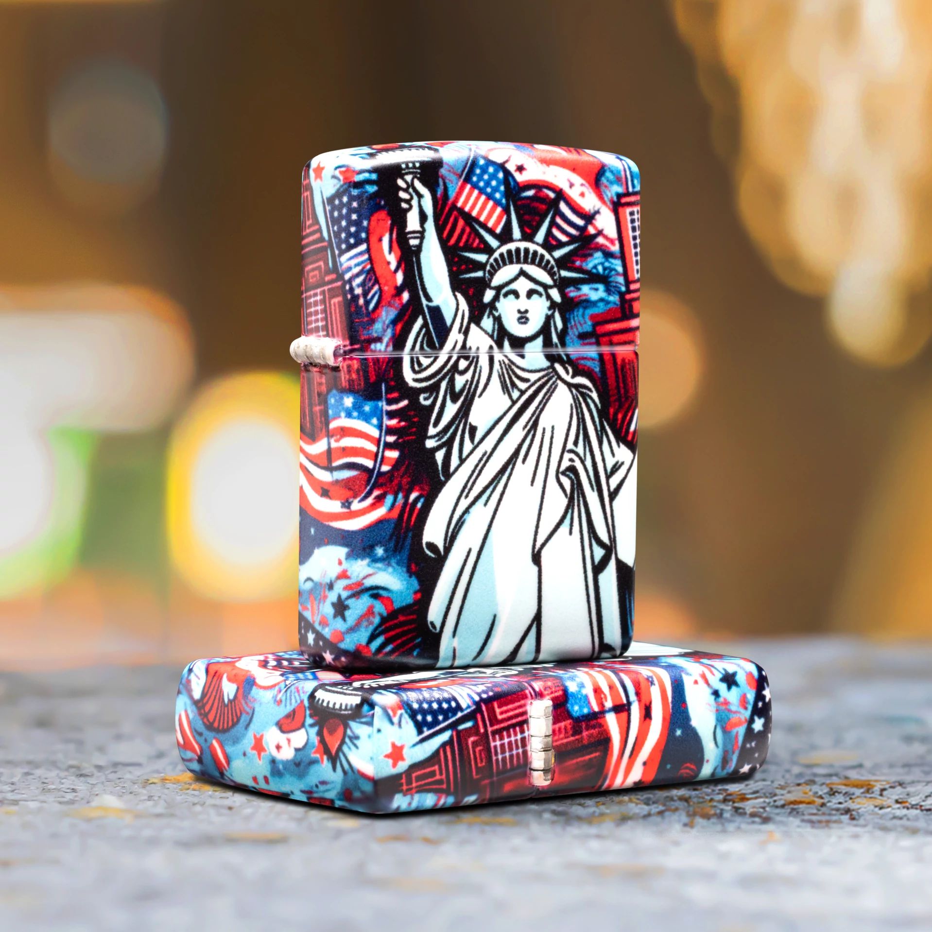 Lifestyle image of two Zippo Lady Liberty Design 540 Matte Windproof Lighters; one lying flat on a concrete surface showing the top of the lid and hinge side of the lighter while the other lighter is standing on top of the lighter that is lying flat, showing the front and hinge side of the lighter with a blurred background. 

