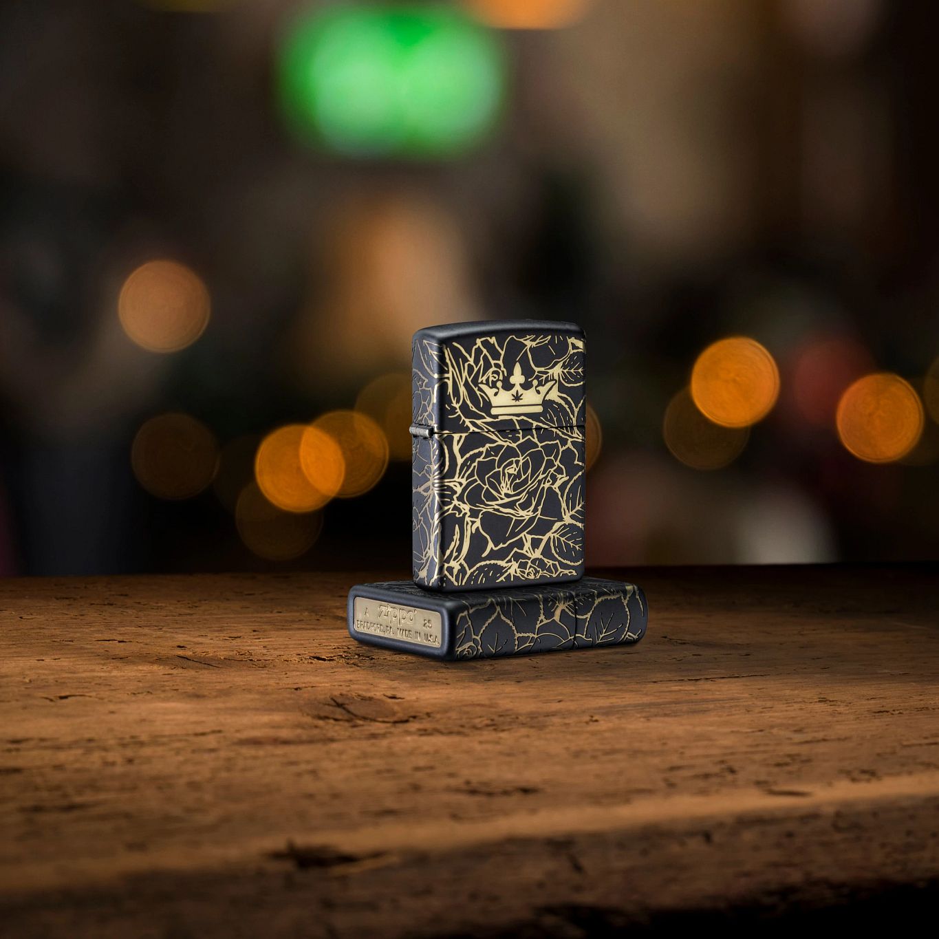 Lifestyle image of two Zippo Queen of Bud 360° Roses Design Black Matte Windproof Lighters; one lying flat on a rustic wooden bar showing the bottom stamp and non-hinge side of the lighter while the other lighter is standing on top of the lighter that is lying flat showing the front of the lighter. 
