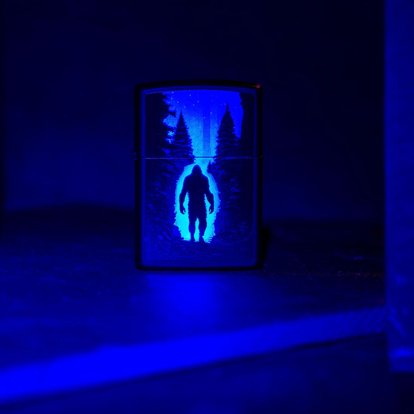 Lifestyle image of Zippo Ultra Squatch Design Black Matte Windproof Lighter standing on a dark surface under a black light. 

