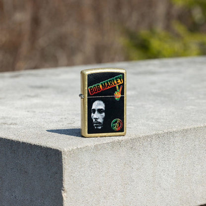 Lifestyle image of Zippo Bob Marley Peace Design Street Brass Windproof Lighter standing on a concrete surface showing the front of the lighter with an out of focus nature background. 

