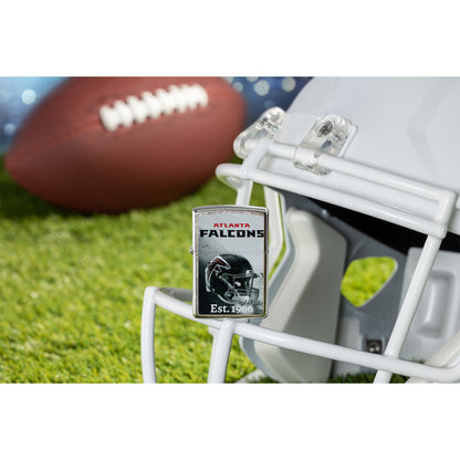 Lifestyle image of Zippo NFL Atlanta Falcons Helmet Design Street Chrome Windproof Lighter standing on the facemask of a white helmet showing the front of the lighter with a football lying on a football field as the background.

