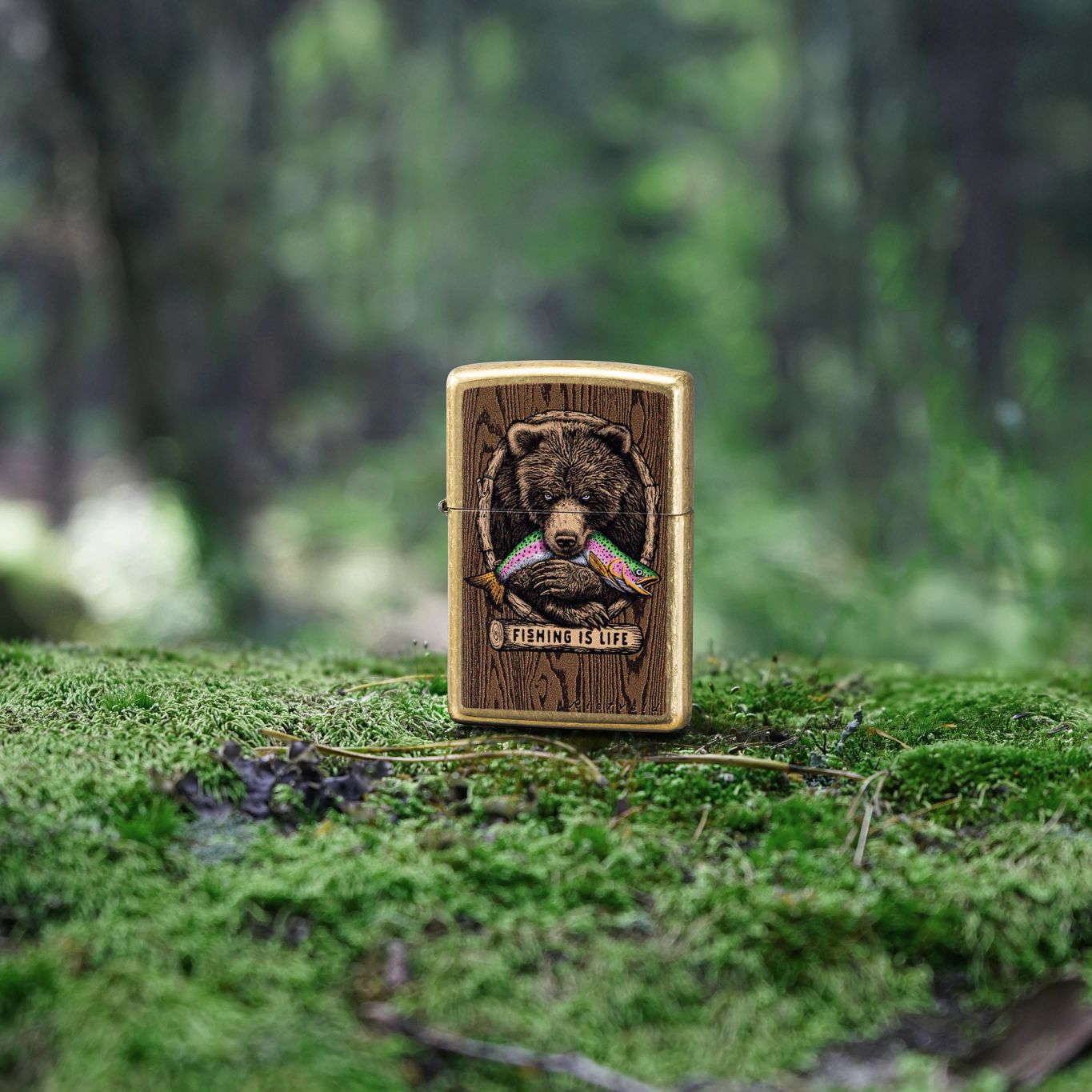 Lifestyle image of Zippo Rietveld Fishing is Life Design Street Brass Windproof Lighter standing on mossy ground showing the front of the lighter with the forest as the background. 

