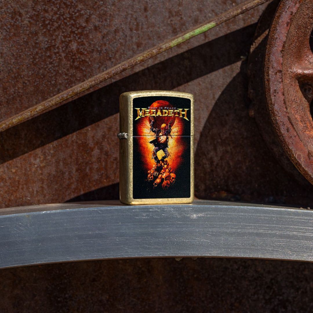 Lifestyle image of Zippo Megadeth Rust In Peace Design Street Brass Windproof Lighter standing in front of rusty gears while showing the front of the lighter. 

