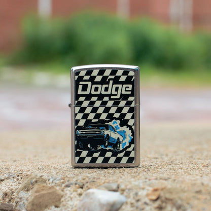 Lifestyle image of Zippo Dodge Dart Design Street Chrome Windproof Lighter standing on concrete showing the front of the lighter with an out of focus background with green bushes and a building.

