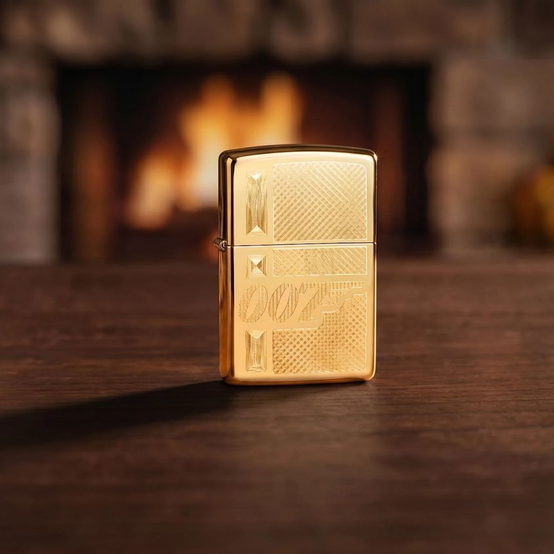 Lifestyle image of Zippo James Bond 007™ Gingham Design High Polish Brass Windproof Lighter standing on a wooden table in front of a fireplace showing the front of the lighter. 

