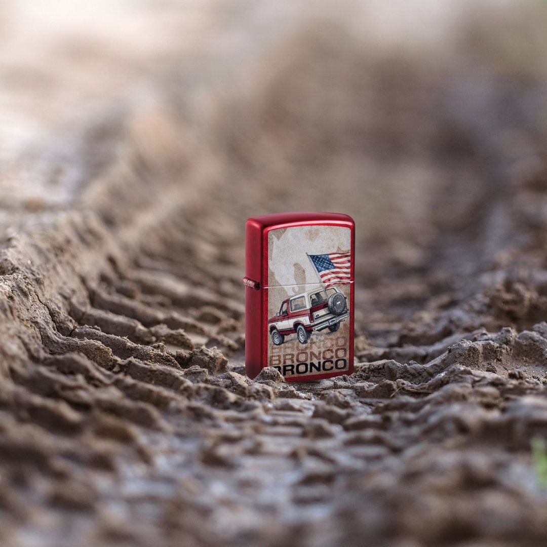 Lifestyle image of Zippo Ford Bronco Flag Design Candy Apple Red Windproof Lighter standing in muddy tire tracks showing the front and hinge side of the lighter. 

