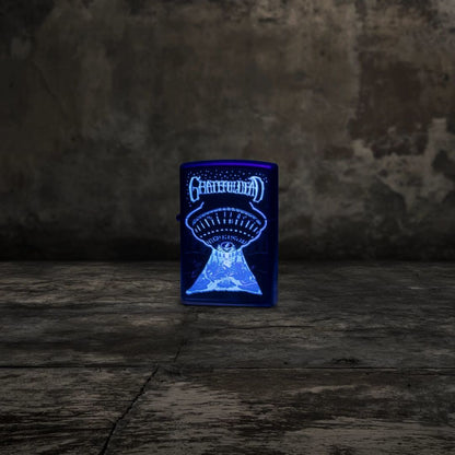 Lifestyle image of Zippo Grateful Dead Black Light UFO Design Black Matte Windproof Lighter standing on a concrete floor showing the front of the lighter while glowing under a black light with a concrete wall as the background. 

