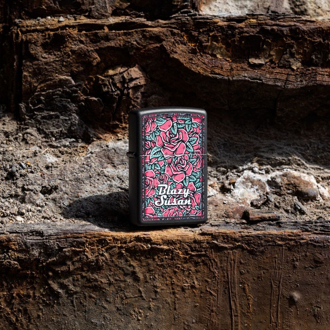 Lifestyle image of Zippo Blazy Susan Roses Design Black Matte Windproof Lighter standing on a rocky surface while showing the front of the lighter. 

