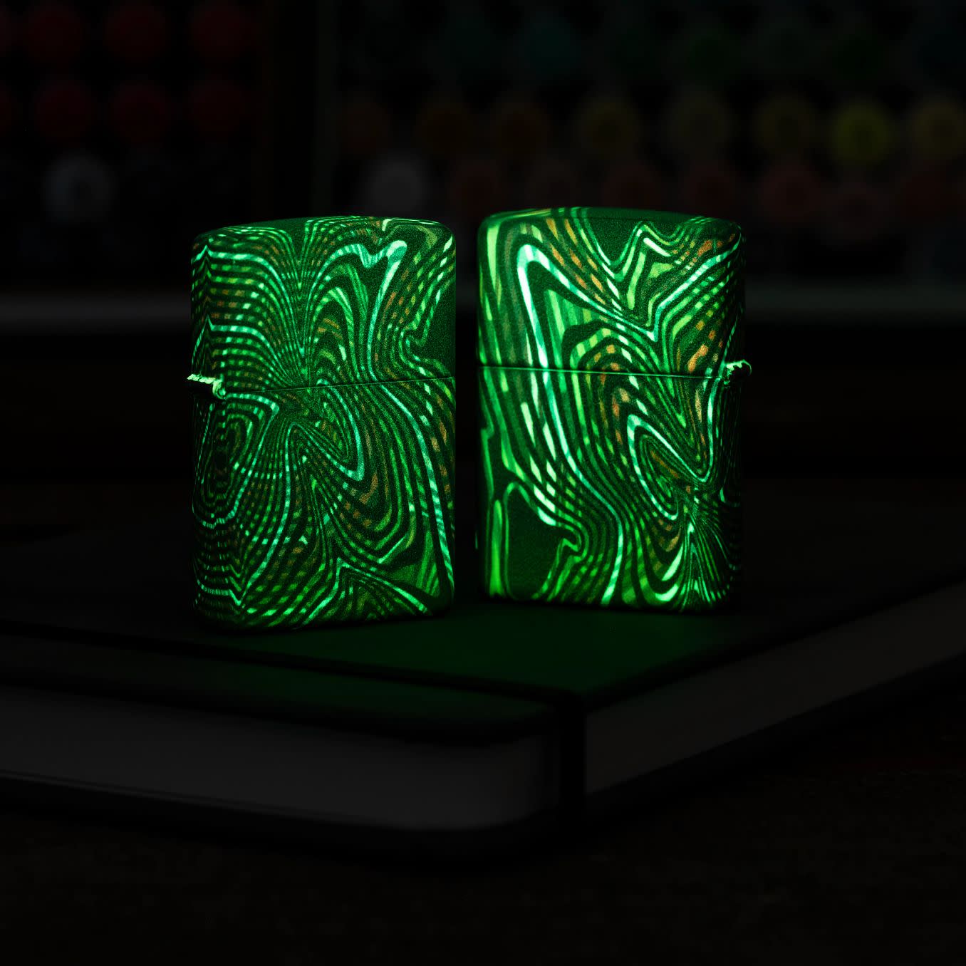 Lifestyle image of two Zippo Glowing Illusion Design Glow in the Dark Windproof Lighters, one showing the front of the lighter and the other showing the back, glowing in the dark.