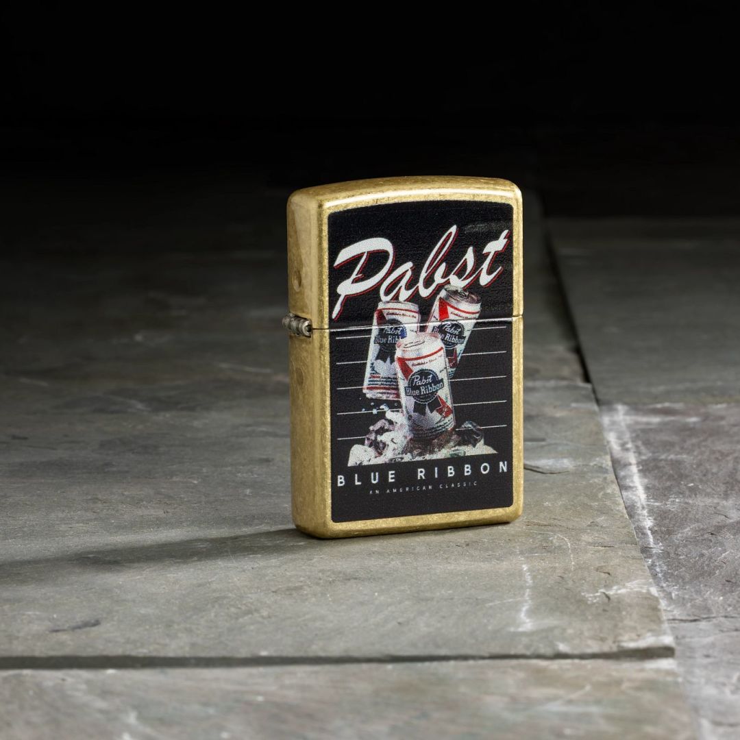 Lifestyle image of Zippo Pabst Blue Ribbon® American Classic Design Street Brass Windproof Lighter standing on tile flooring showing the front and hinge side of the lighter. 

