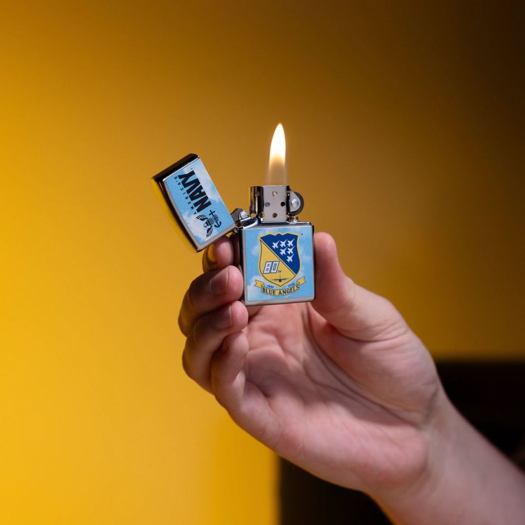 Lifestyle image of Zippo United States Navy® Blue Angels Design Street Chrome Windproof Lighter lit in hand showing the front of the lighter with a yellow wall as the background. 

