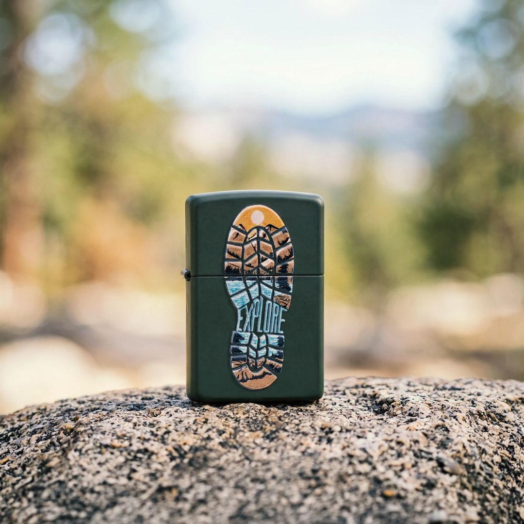 Lifestyle image of Zippo Step Out Design Green Matte Windproof Lighter standing on a rock showing the front of the lighter with an out of focus nature background. 

