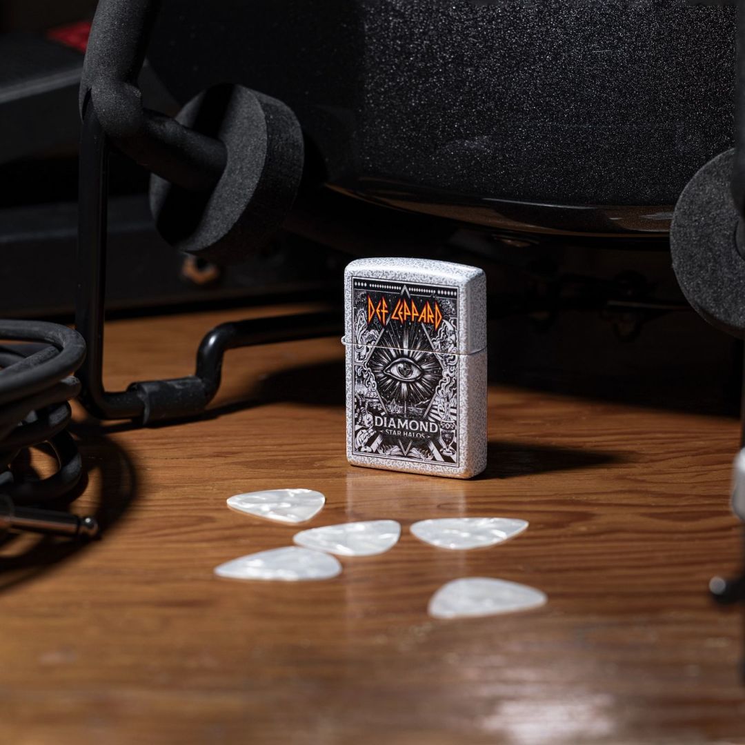 Lifestyle image of Zippo Def Leppard Diamond Star Halos Design Glacier Windproof Lighter standing on a table next to some white guitar picks and some other music equipment while showing the front and non-hinge side of the lighter. 

