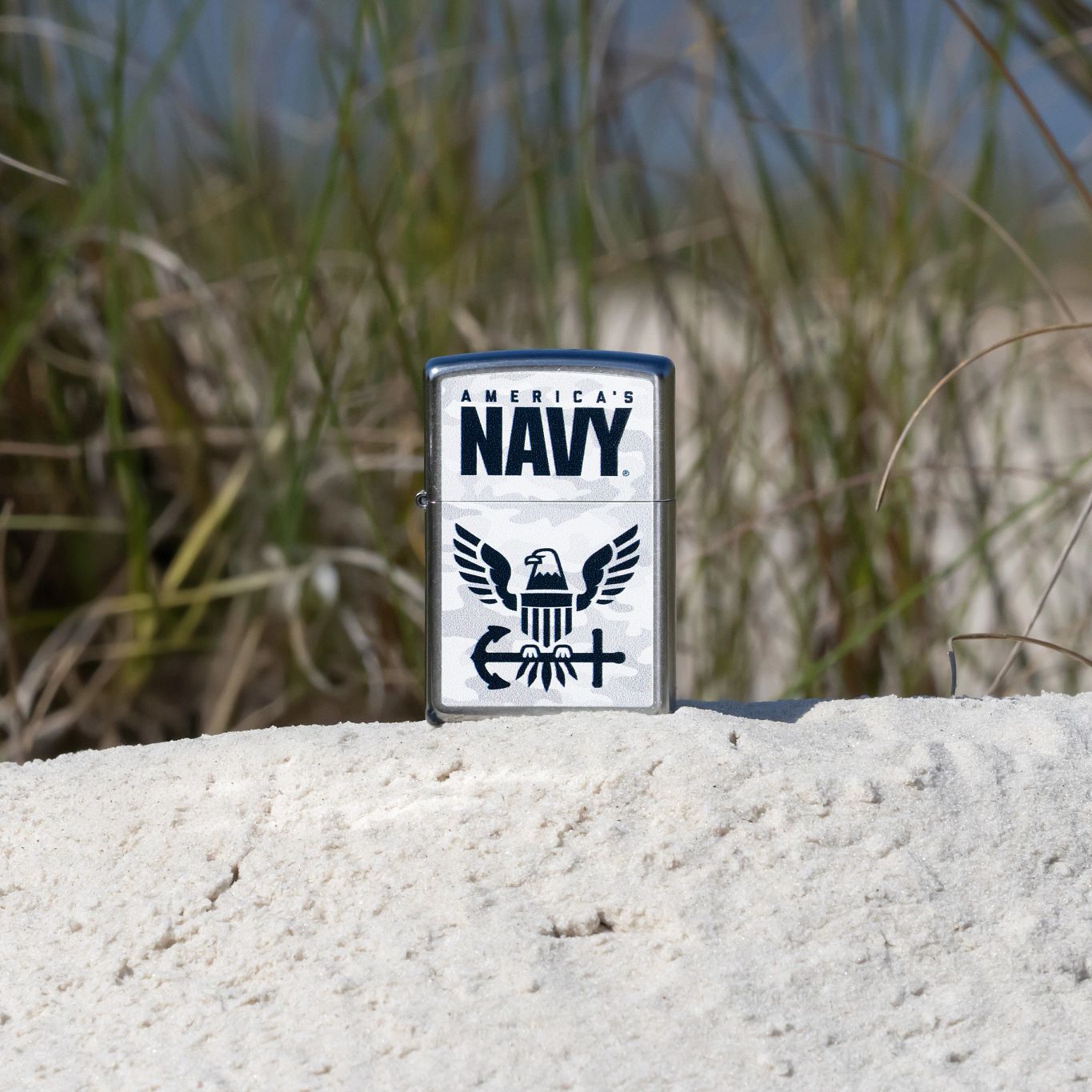 Lifestyle image of Zippo United States Navy® Camo Logo Design Street Chrome Windproof Lighter standing on a grassy sand dune showing the front of the lighter. 

