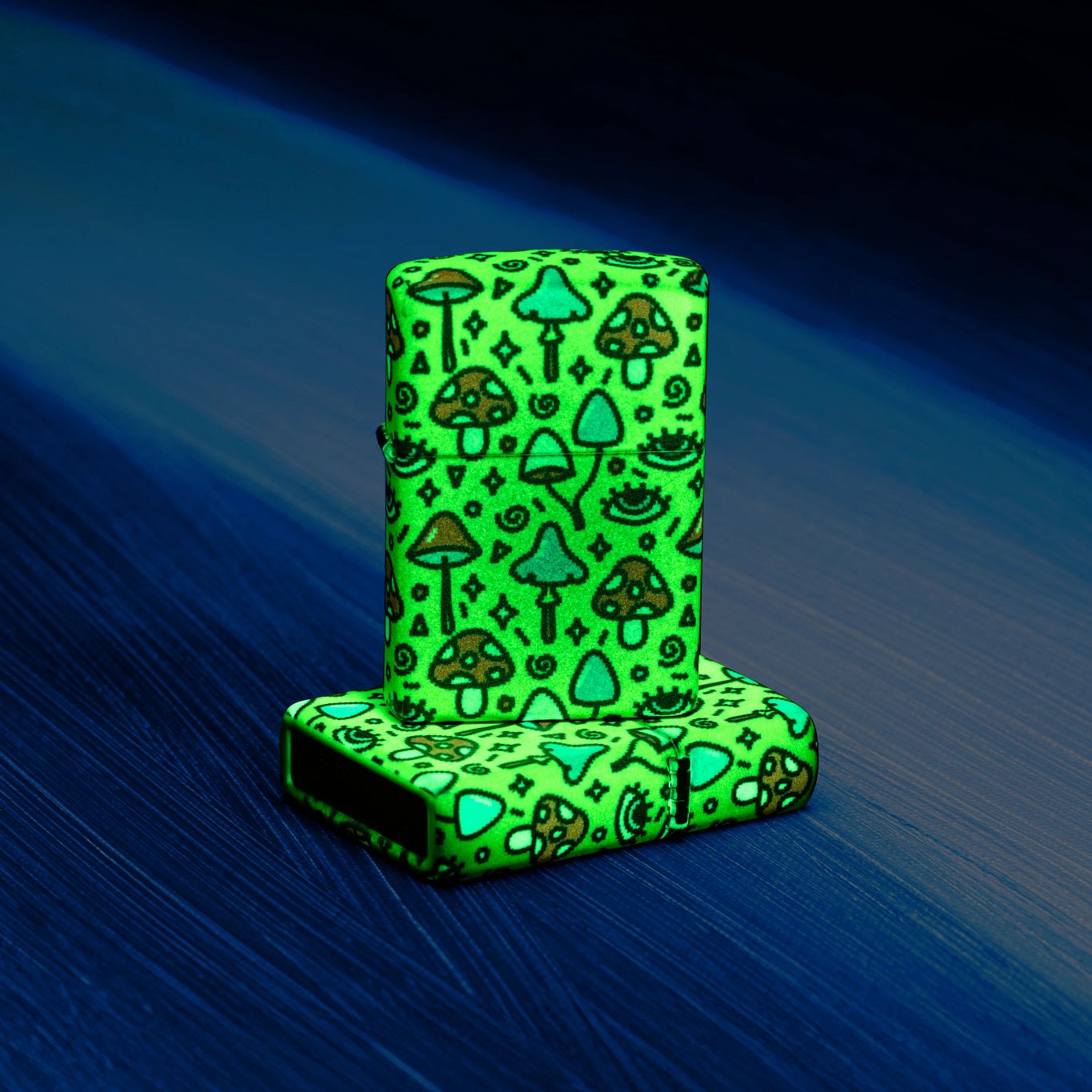 Lifestyle image of two Zippo Eye Spy Design Glow-in-the-Dark Green Windproof Lighters; one lying flat on a wood grain surface showing the hinge side of the lighter, while the other lighter is standing on top of the flat lighter, both glowing in the dark.
