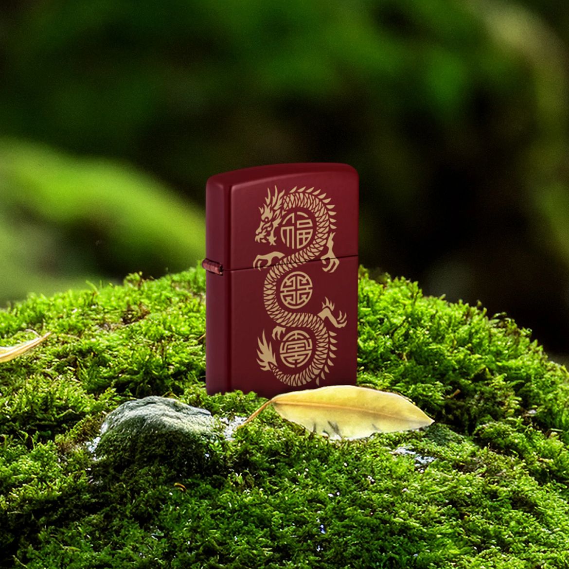 Lifestyle image of Zippo Lucky Dragon Design Merlot Windproof Lighter standing on moss covered ground next to two yellow leaves with a blurred mossy background.

