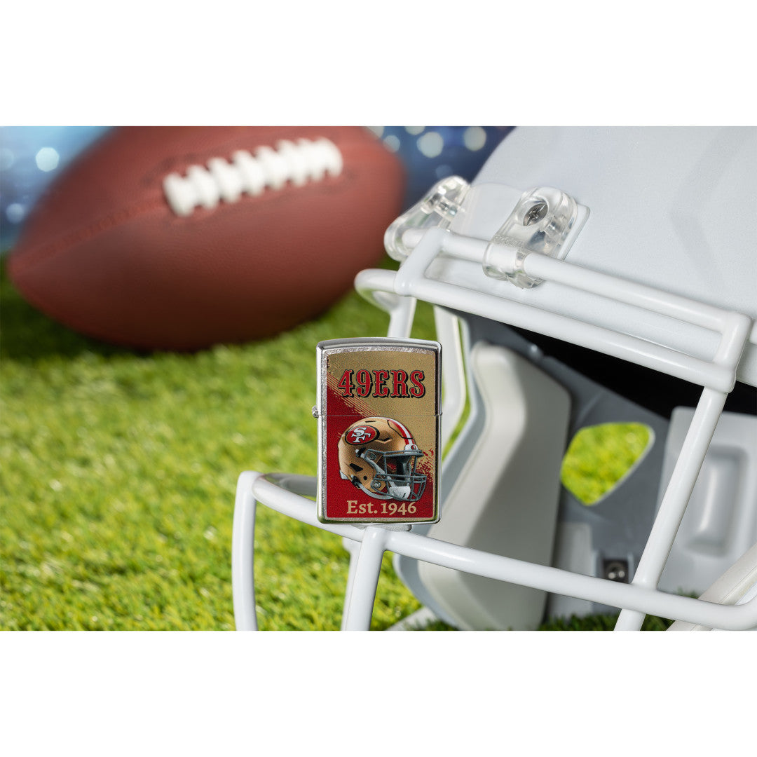 Lifestyle image of Zippo NFL San Francisco 49ers Helmet Design Street Chrome Windproof Lighter standing on the facemask of a white helmet showing the front of the lighter with a football lying on a football field as the background.

