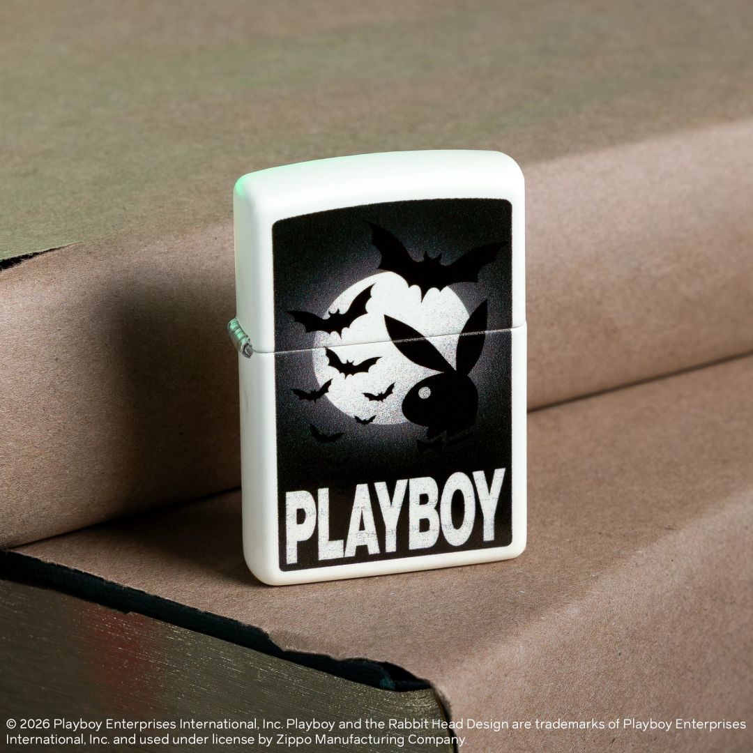 Lifestyle image of Zippo Playboy Glowing Bats Design Glow In The Dark Windproof Lighter standing on a book covered in lighter brown paper next to another book covered in light brown paper while showing the front and hinge side of the lighter. 

