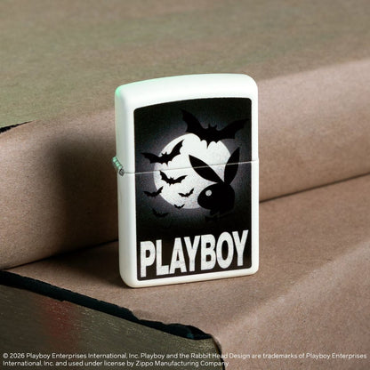 Lifestyle image of Zippo Playboy Glowing Bats Design Glow In The Dark Windproof Lighter standing on a book covered in lighter brown paper next to another book covered in light brown paper while showing the front and hinge side of the lighter. 

