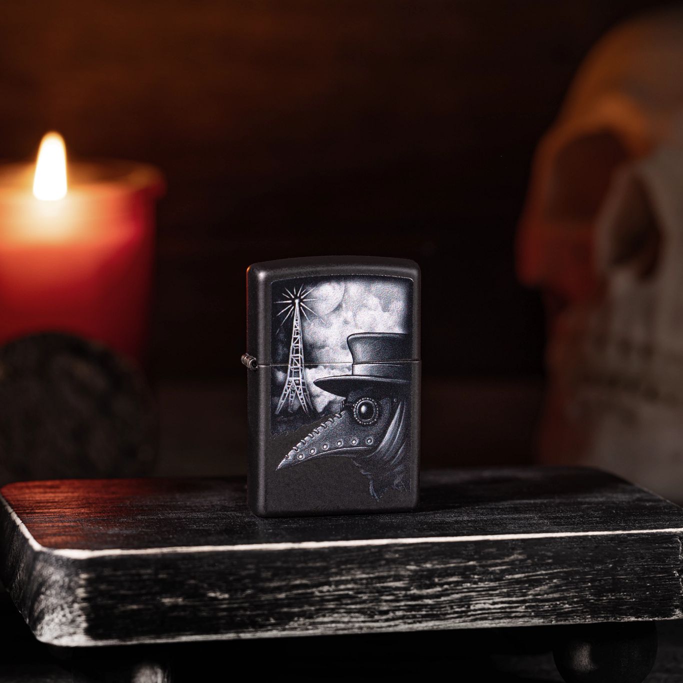 Lifestyle image of Plague of Disinformation Black Matte Windproof Lighter standing on a dark wooden stool with a lit candle and skull in the background. 

