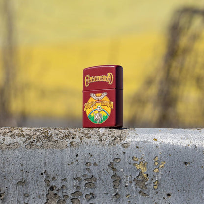 Lifestyle image of Zippo Grateful Dead Sunshine Daydream Design Merlot Windproof Lighter standing at a 3/4 angle on the ledge of a grey painted concrete structure showing the front and non-hinge side of the lighter with a yellow and grey background. 

