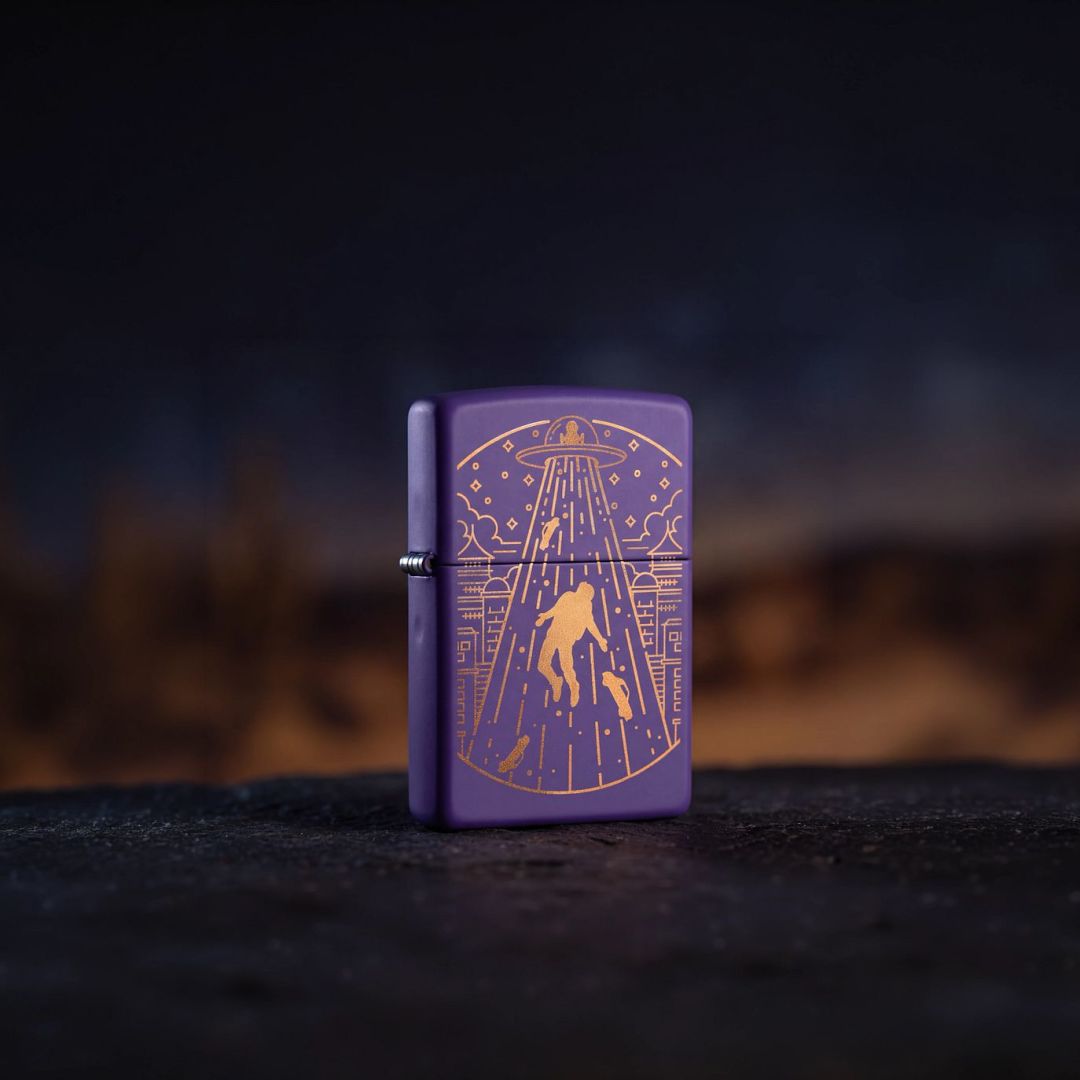 Lifestyle image of Zippo Beam Me Up Design Purple Matte Windproof Lighter standing on a black surface showing the front and hinge side of the lighter with an out of focus dark background. 

