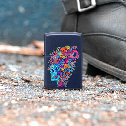 Lifestyle image of Zippo Skull Reef Design Navy Matte Windproof Lighter standing in gravel next to a blue wall and a black leather boot, while showing the front of the lighter. 

