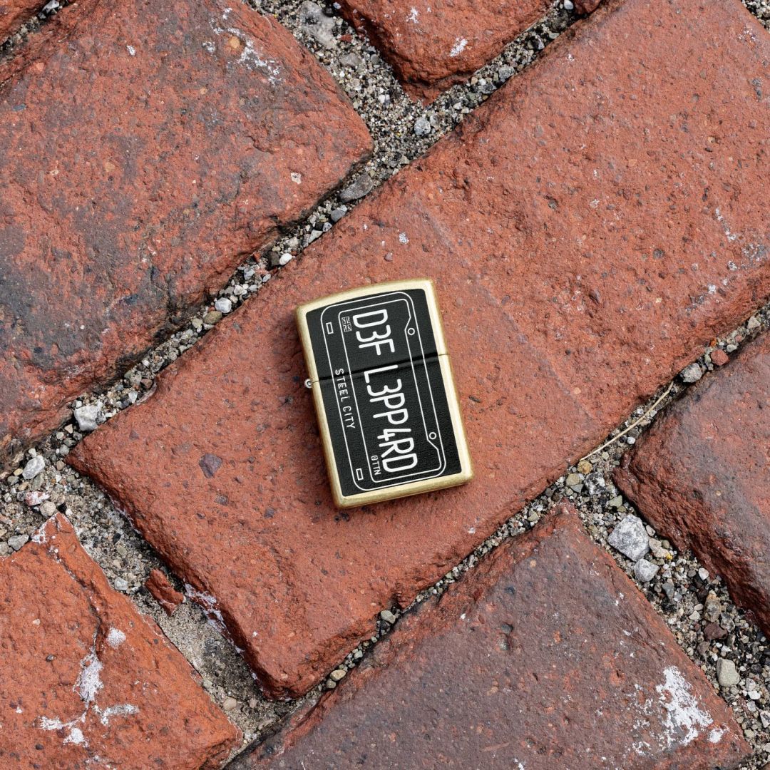 Lifestyle image of Zippo Def Leppard Steel City Design Street Brass Windproof Lighter lying on a brick walkway showing the front of the lighter. 

